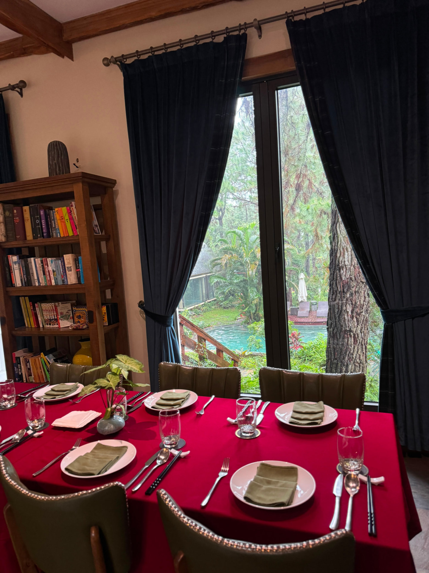 Dinner table laid for six beside a window looking onto a pine-shaded pool